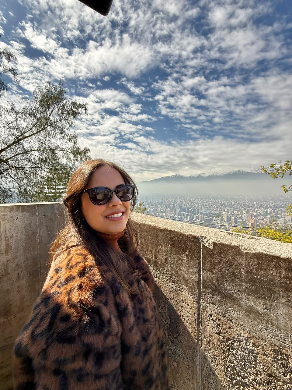 Carol Regazi de óculos escuros e casaco em viagem, com cidade e montanhas ao fundo